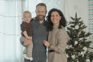A joyful family with a baby poses next to a decorated Christmas tree indoors.