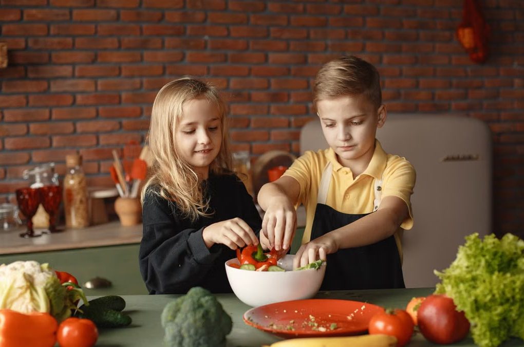 Cheerful girl helping little boy in apron preparing salad while standing in kitchen near table with mix of fresh vegetables behind brick wall