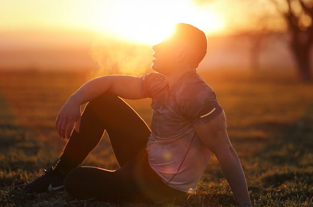 Muscular man sits in rural field at sunset, exhaling visible breath.